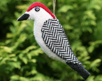 May include: A fabric bird ornament featuring a cream-colored body, a bright red crown, and a black pointed beak. The wing displays a black and white chevron pattern. A small black button serves as the eye, and a thin cord is attached for hanging.