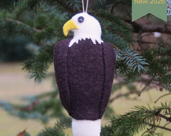 May include: A felt bald eagle ornament featuring a white head, a dark brown body, a white tail, and a yellow beak. A small dark blue button serves as the eye. A silver string is attached to the top. A green tag displays the text New 2026.