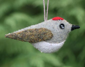 May include: A felt bird ornament featuring a light gray body, a white underbelly, and a textured brown wing. The bird has a small red patch on its head, a black button eye, and a pointed black beak. A brown and white striped cord is attached.