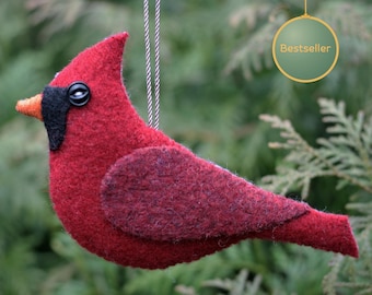 May include: A red felt cardinal bird ornament with a black face patch, an orange beak, and a black button eye. The bird is suspended by a silver cord and features a circular tag in the upper right corner with the text Bestseller.