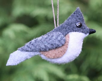 May include: A felt bird ornament featuring a dark gray crest, a light gray wing, a tan side patch, and a white chest. The bird has a small black button eye, a black beak, and is suspended by a thin, brown and white twisted cord for hanging.