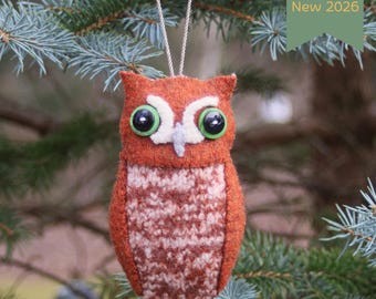 May include: An orange and brown felt owl ornament with large green button eyes and a white felt face patch. The owl features a textured knit-style belly and hangs from a silver cord. A green tag in the corner displays the text New 2026.