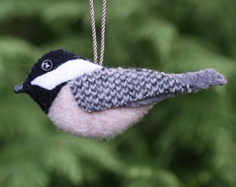May include: A felted bird ornament featuring a black head with a white cheek patch, a grey knitted-texture wing, and a light beige underbelly. The bird has a small black button eye and is suspended by a thin brown and white twisted cord.