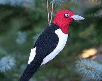 May include: A felt ornament shaped like a red-headed woodpecker with a bright red head, a black back and tail, and a white chest. The bird features a small black button for an eye and a grey beak, suspended by a thin, twisted metallic cord.