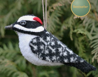 May include: A felted woodpecker ornament featuring a black and white patterned body, a white chest, a black head with a white stripe, and a small red patch on the crown. The bird has a black button eye and hangs from a brown and white cord.