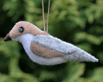 May include: A felt bird ornament featuring a tan head, a white chest and underbelly, and light gray wings and tail. The bird has a small dark brown beak and a black button eye. A thin, braided cord is attached to the top for hanging purposes.