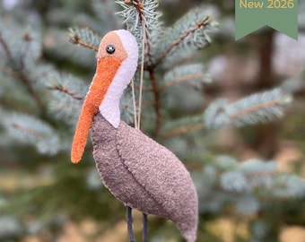 May include: A felt pelican ornament featuring a brown body, a white neck, and a textured orange beak with a small black button eye. The bird is suspended by a thin string and includes a green tag with the text New 2026.