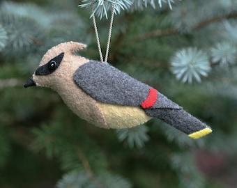 May include: A felt bird ornament featuring a tan head with a black eye patch and crest, a grey back and wing, a red band on the wing, a pale yellow underbelly, and a grey tail with a yellow tip, suspended by a thin silver cord for hanging.