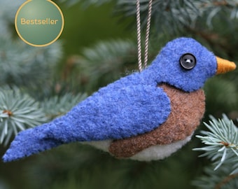 May include: A felt bird ornament featuring a blue head, wings, and tail, a brown chest, and a white belly. The bird has a small black button eye, an orange beak, and a thin string loop for hanging. The ornament is textured and soft in appearance.