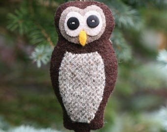 May include: A felt owl ornament featuring a dark brown body, a light gray textured belly, and a light gray face patch. The owl has two black button eyes, a small yellow felt beak, and a thin silver hanging string attached to the top of its head.