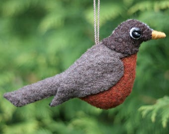 May include: A felted bird ornament featuring a dark brown head, a grey back and tail, and a vibrant orange-red chest. The bird has a small yellow beak and a single black bead eye. A thin silver cord is attached to the top for hanging purposes.