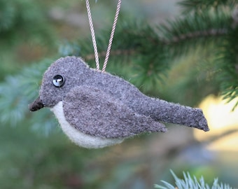 May include: A felt bird ornament featuring a dark gray back and wings with a light cream-colored belly. The bird has a small black button eye and a dark brown beak. A thin, twisted brown and white cord is attached to the top for hanging purposes.