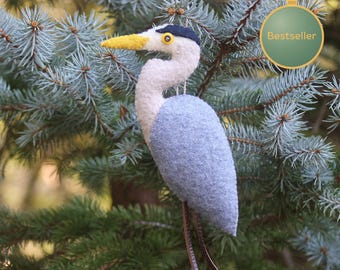 May include: A felted heron ornament featuring a light blue body, a white neck, a dark blue head crest, and a yellow beak. The bird has a small black eye and two thin brown legs. A circular tag with the text Bestseller is attached.