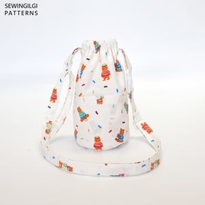 May include: White drawstring bag with a long strap. The bag has a white background with a pattern of brown bears wearing sweaters and colorful sprinkles. The bag has a small pocket on the front.