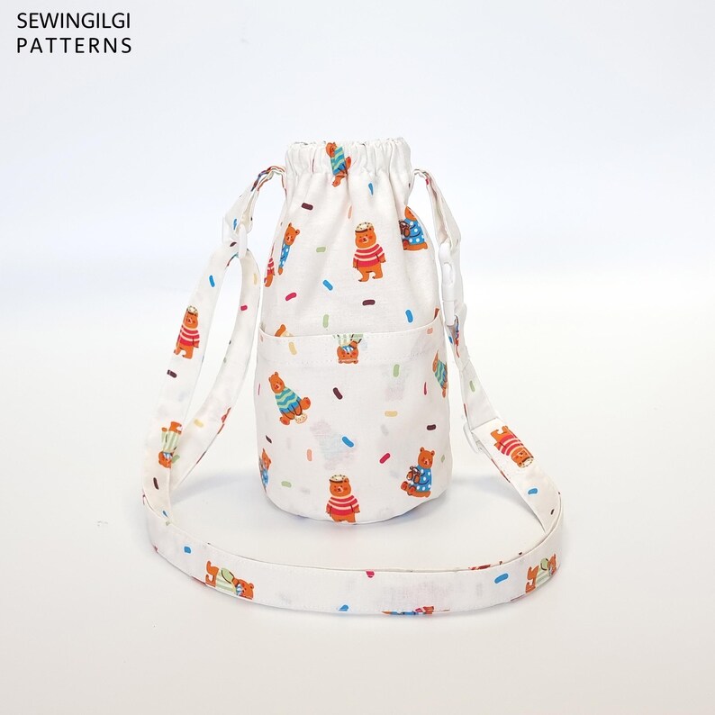 May include: White drawstring bag with a long strap. The bag has a white background with a pattern of brown bears wearing sweaters and colorful sprinkles. The bag has a small pocket on the front.