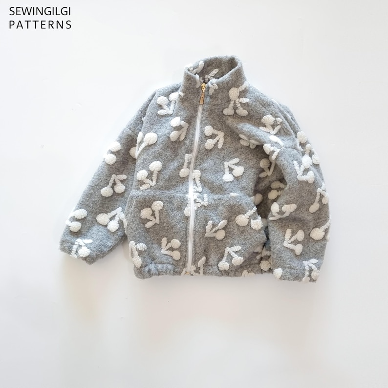 May include: A grey fleece zip-up jacket featuring a white cherry pattern, a high collar, and two front pockets. The text SEWINGILGI PATTERNS is visible in the top left corner. The jacket has a white zip closure and long sleeves.