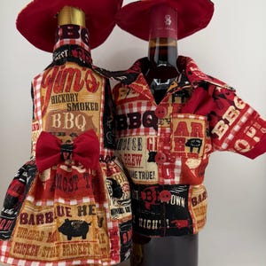 May include: Two wine bottles dressed in BBQ-themed outfits. One bottle wears a red and white checkered dress with a bow, the other a matching shirt and hat. Both have red hats and are decorated with BBQ-related text and images.