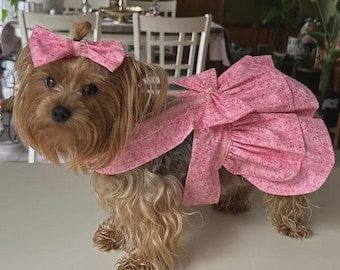 Peut inclure: Un chien Yorkshire Terrier portant une robe rose et un nœud assorti dans ses cheveux. La robe présente un motif floral et un grand nœud dans le dos. Le chien a des poils bruns et fauves.