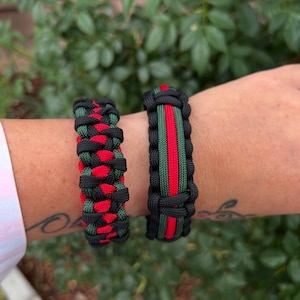 May include: Two black paracord bracelets. One features a red and green pattern, the other has a red and green stripe down the center. Both are handmade and suitable for everyday wear.