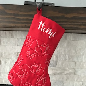 May include: A red Christmas stocking with white embroidered paw prints and the name "Hemi" in white script. The stocking is hanging from a metal hook, with a wooden mantel and a white stone wall in the background.