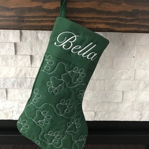 May include: A green Christmas stocking with white embroidered paw prints and the name "Bella" in cursive. The stocking is hanging from a metal hook on a wooden mantel, with a white brick background.
