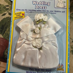 May include: A white satin wedding dress for Webkinz pets, packaged in a blue and yellow card. The dress features white rose accents and the text "Wedding Dress". The package includes a feature code.