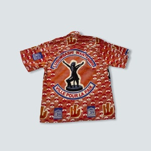 May include: A red and white short-sleeved shirt with a large circular design on the back. The design features a black silhouette of a person with arms raised, surrounded by the text "Lubumbashi Wantaanshi Ville Pour La Paix" and the UNESCO logo.