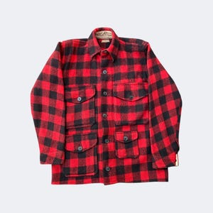 May include: A red and black plaid wool jacket with four front pockets and a collared neckline. The jacket has black buttons and a classic lumberjack style. The fabric appears to be thick and warm, suitable for cold weather.