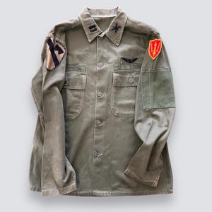 Puede incluir: Una camisa militar verde con varios parches, incluyendo un parche blanco y negro con el número "1", un parche rojo y dorado con un tridente y un parche negro con el texto "U.S. ARMY".