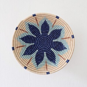 May include: A round woven basket with a floral design. The basket features a central navy blue flower pattern surrounded by light blue and tan concentric circles. Brown and navy blue accents are incorporated throughout the design.