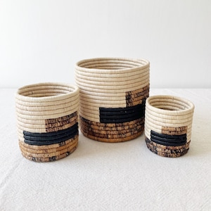 Può includere: Tre cesti intrecciati di varie dimensioni. I cesti sono cilindrici con un colore beige naturale, accentuati da un motivo geometrico marrone scuro e nero alla base. Il cesto più grande è alto circa 25 cm.