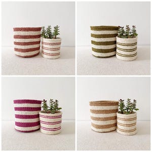 May include: A collection of four sets of woven baskets, each featuring a large and small cylindrical basket with horizontal stripes in terracotta, olive green, magenta, and tan, paired with a small green succulent plant inside each.