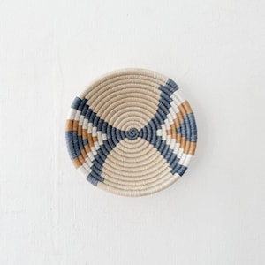 May include: A round, shallow woven basket featuring a coiled design with alternating patterns of natural beige, slate blue, and warm tan fibers. The circular construction radiates from a central point, creating a symmetrical geometric motif.