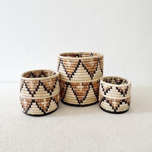 May include: Three woven baskets with a geometric pattern in shades of brown, black, and white. The baskets are nested inside each other, with the largest basket at the bottom and the smallest basket at the top.
