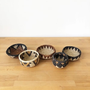 Può includere: Cinque piccoli cestini rotondi intrecciati con motivi geometrici nei toni del marrone, nero e beige. I cestini sono disposti su una superficie di legno chiaro, mostrando le loro diverse dimensioni e design. Le trame e i colori naturali creano un'estetica rustica.