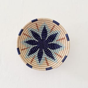 May include: A circular woven basket with a starburst pattern. The basket has alternating bands of beige, light blue, brown, and navy blue. The centre of the basket is navy blue, with the design radiating outwards.