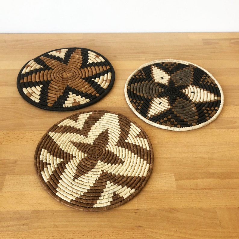 May include: Three round woven placemats with a brown, black, and white geometric pattern. The placemats are made of natural materials and have a rustic, handmade look.