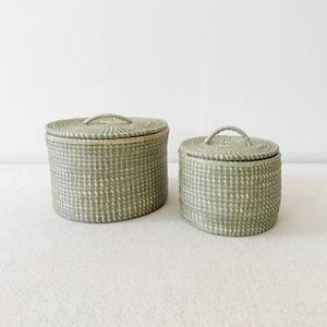 May include: Two woven storage baskets with lids. The baskets are cylindrical, with a light green and white colour scheme. Each basket has a handle on the lid. The larger basket is approximately 25 cm in diameter, and the smaller basket is about 18 cm in diameter.