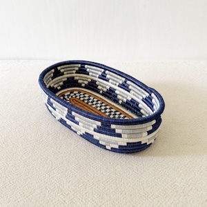 May include: Oval woven basket with a geometric pattern in navy blue, white, and gray. The interior features a brown and black checkered design. This decorative basket is ideal for storage or display.