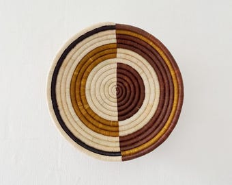 Puede incluir: Una cesta redonda tejida con un diseño dividido. Una mitad presenta círculos concéntricos crema, dorados y marrones, la otra mitad tiene tonos marrones y dorados. Una banda negra bordea la sección crema. La cesta probablemente se utiliza para decoración o almacenamiento.