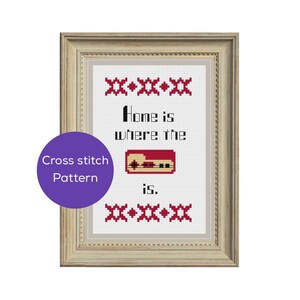 Könnte beinhalten: Gerahmtes Kreuzstich-Kunstwerk mit dem Text "Home is where the is." Das Design zeigt einen roten und schwarzen Videospiel-Controller. Ein violetter Kreis mit "Cross stitch Pattern" befindet sich unten links. Der Rahmen ist hellbraun.