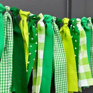 May include: A green fabric garland with a variety of green ribbons. Some ribbons have white clover patterns, while others feature gingham patterns. The garland is knotted at the top, creating a decorative and celebratory appearance.