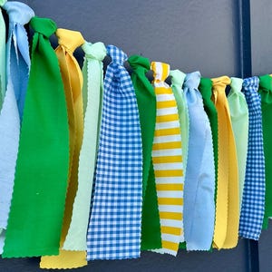 Puede incluir: Una colorida guirnalda de tela con cintas anudadas en varios tonos de verde, azul, amarillo y blanco. Algunas cintas tienen patrones de cuadros o rayas. Ideal para decoraciones festivas.