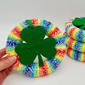 May include: Rainbow-colored, plush St. Patrick's Day wreath with a large green felt shamrock. A stack of three similar wreaths is also visible. The wreaths are made of a soft, textured material.