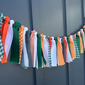Może przedstawiać: Świąteczna girlanda z tkaniny z r&oacute;żnokolorowymi wstążkami. Girlanda zawiera wstążki w kolorze pomarańczowym w białe grochy, zielono-białą kratę vichy, jednolity pomarańczowy, biały i zielony. Girlanda jest zawiązana i wisi na ciemnoszarym tle.