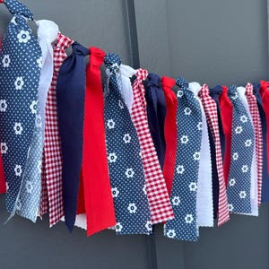 Może przedstawiać: Świąteczna girlanda z tkaniny z naprzemiennymi czerwonymi, białymi i granatowymi wstążkami. Niekt&oacute;re wstążki mają biały kwiatowy wz&oacute;r na granatowym tle, inne są jednolicie czerwone lub granatowe, a niekt&oacute;re są białe lub czerwone w kratkę.