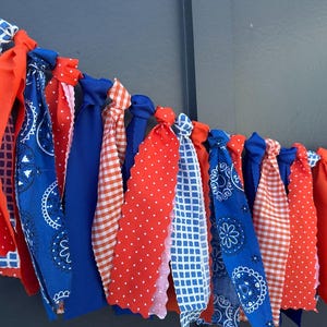 Może przedstawiać: Świąteczna girlanda z naprzemiennymi paskami materiału w kolorach czerwonym, białym i niebieskim. Girlanda zawiera jednolite kolory, grochy, kratę i wzory paisley, tworząc dekoracyjny akcent.