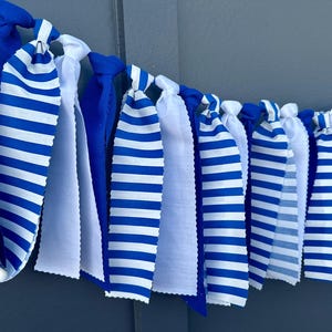 Może przedstawiać: Dekoracyjny baner z naprzemiennymi niebieskimi i białymi paskami materiału. Niekt&oacute;re paski są jednolicie niebieskie, a inne mają niebieskie i białe poziome paski. Baner jest zawiązany u g&oacute;ry.