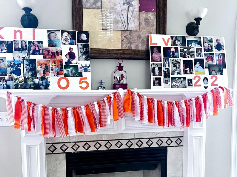 May include: A white mantelpiece decorated with a garland of orange, red, and white fabric strips. Above the mantel are two white canvases with photos and the numbers "05" and "2022" in orange. The canvas on the left has the name "Kni" in red.