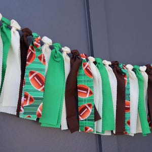 Puede incluir: Una guirnalda de tela con tiras alternas de color crema, marrón oscuro, verde brillante y tela verde estampada con un patrón de balones de fútbol americano marrones y blancos, atadas a lo largo de una cuerda horizontal.